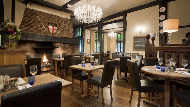 A restaurant with wooden tables and chairs, a brick wall, and a chandelier. - The George Hotel (Cranbrook)