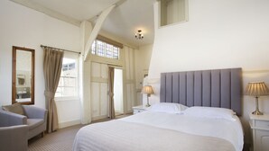 A bedroom with a large bed, two bedside tables with lamps, a chair, a window with curtains, and a mirror on the wall. - The George Hotel (Cranbrook)