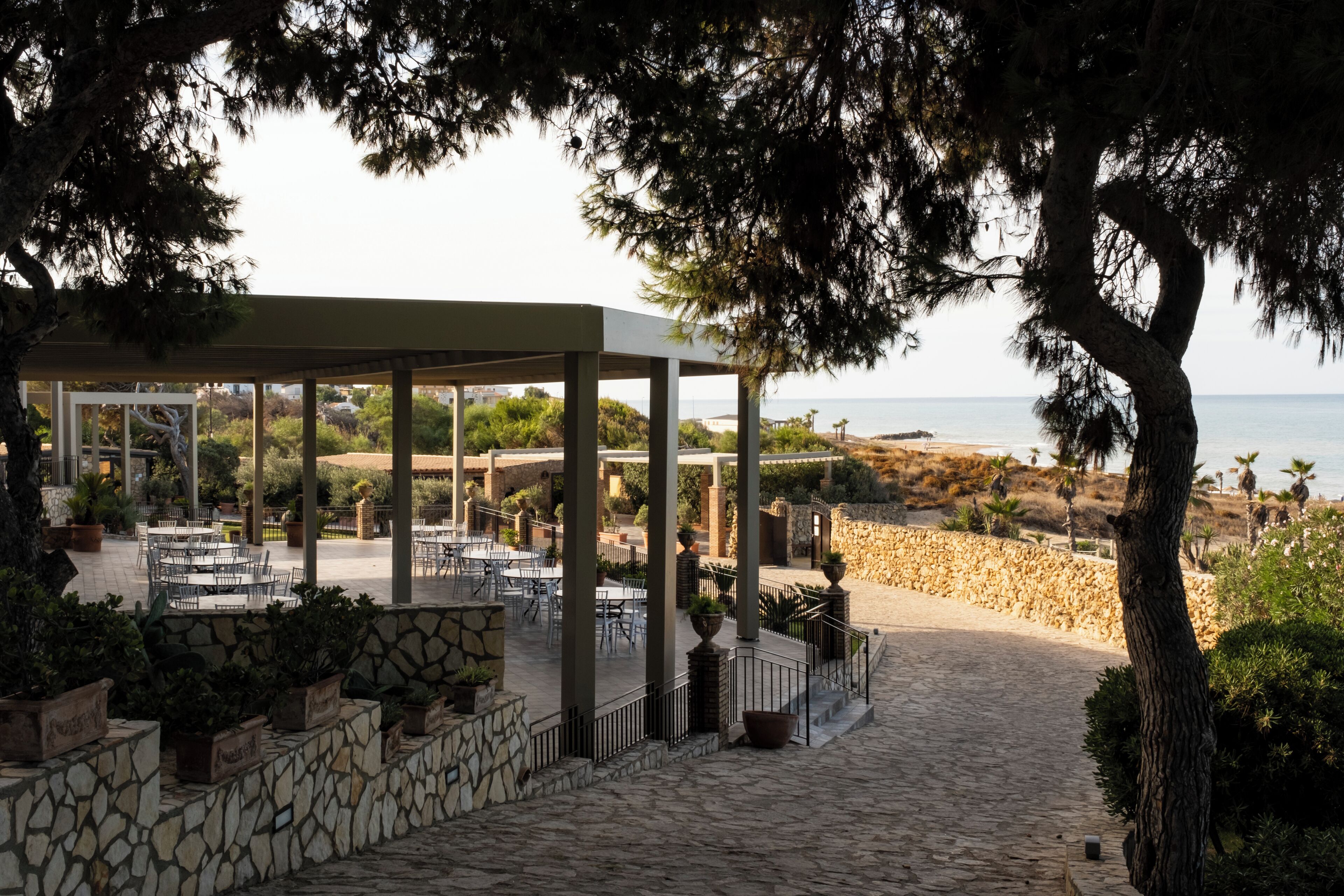A covered outdoor seating area with stone walls and potted plants, overlooking a beach and ocean.