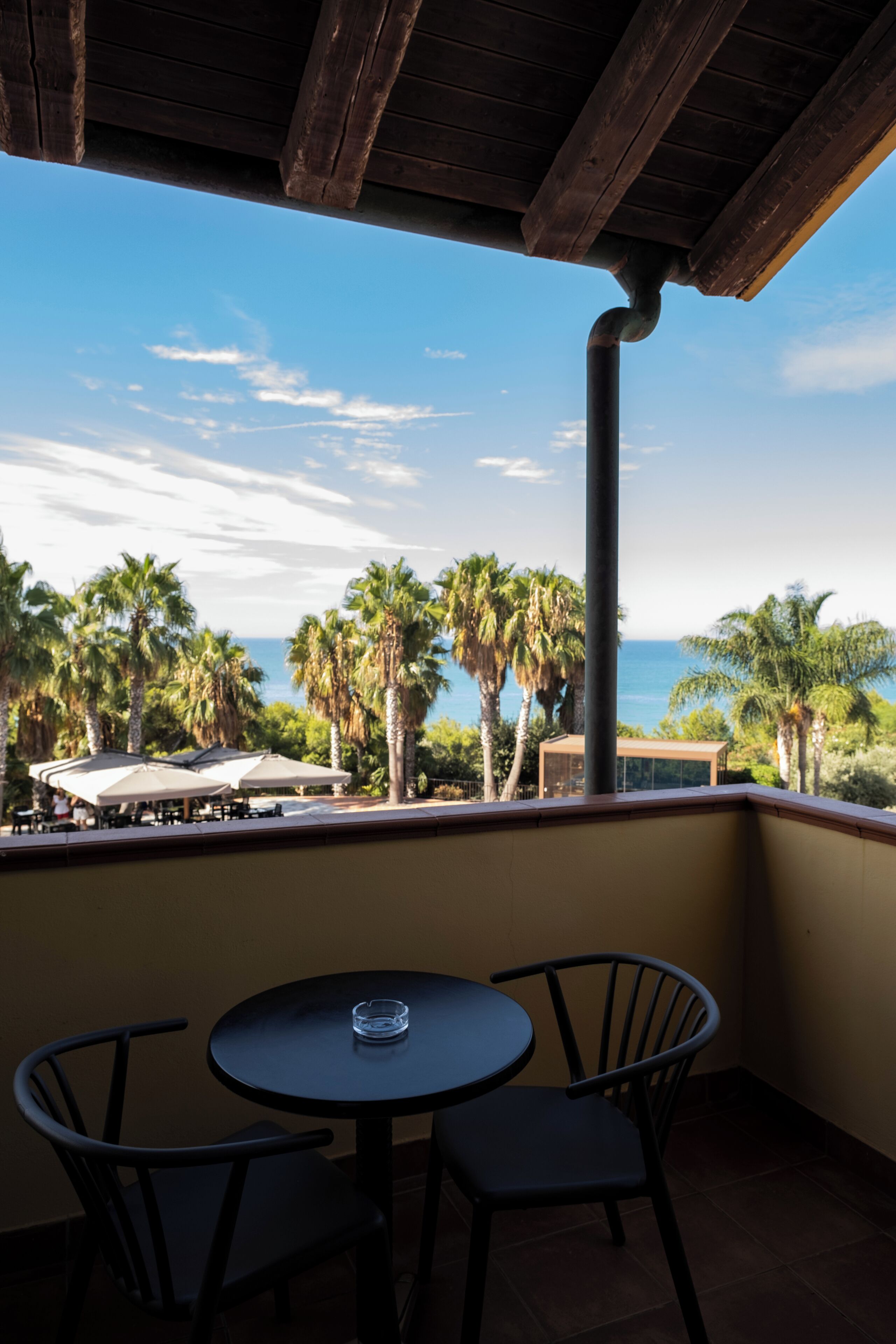 A balcony with a table and chairs, overlooking a beach with palm trees and the ocean.