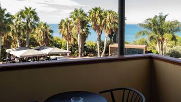 A balcony with a table and chairs, overlooking a beach with palm trees and the ocean.