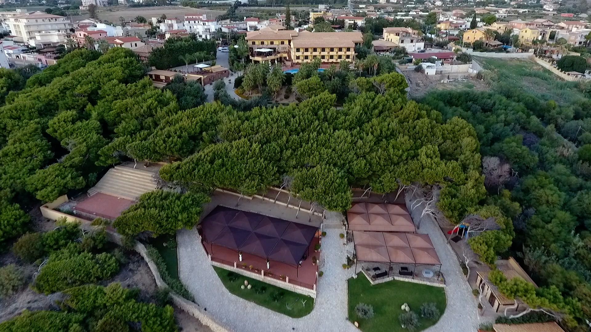 An aerial view of a residential area with a large building surrounded by dense greenery, a paved area with a covered structure, and a clear sky.