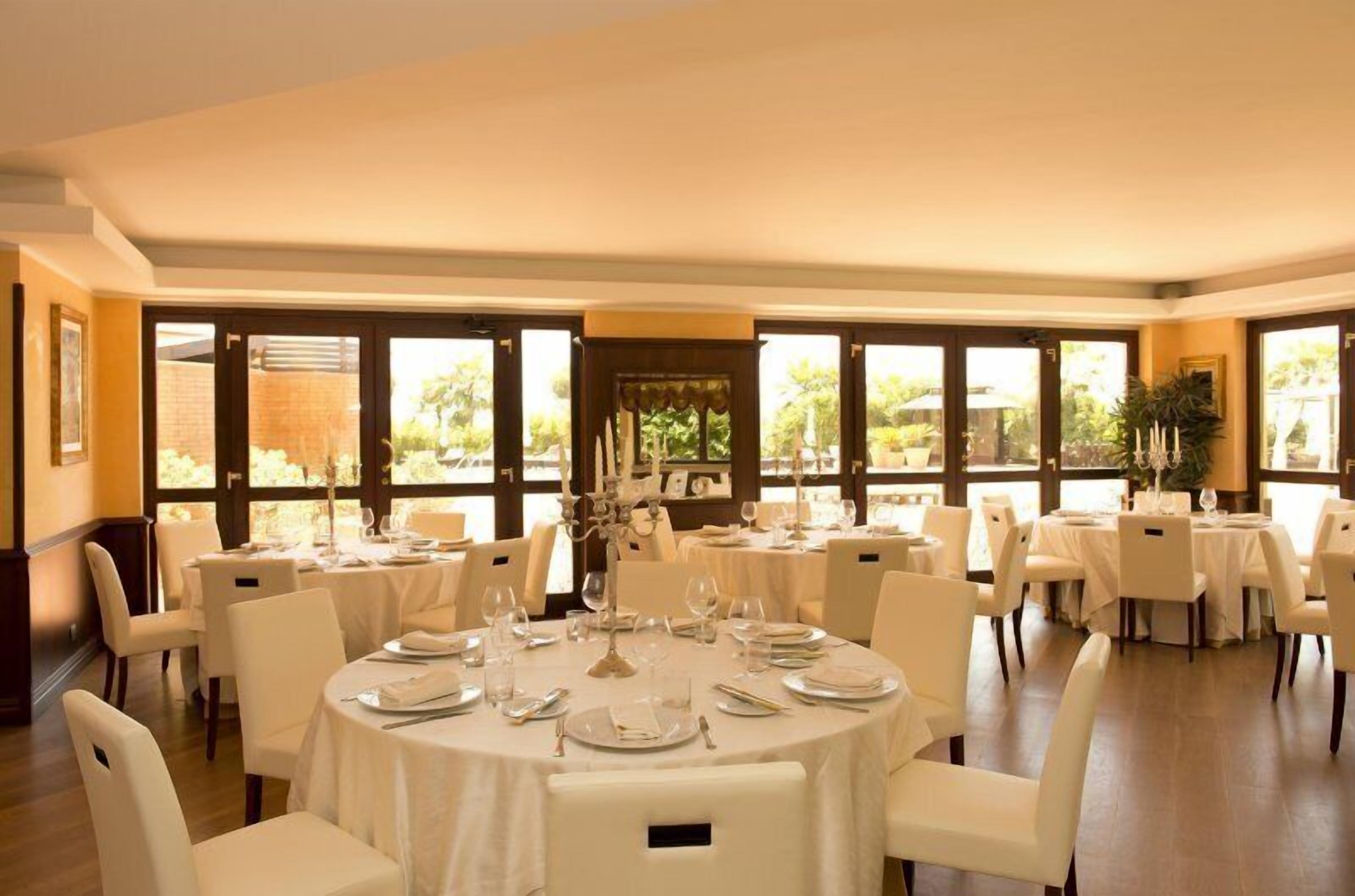 Un espace repas avec des tables rondes dressées pour le service, de grandes fenêtres et une vue sur l’extérieur.