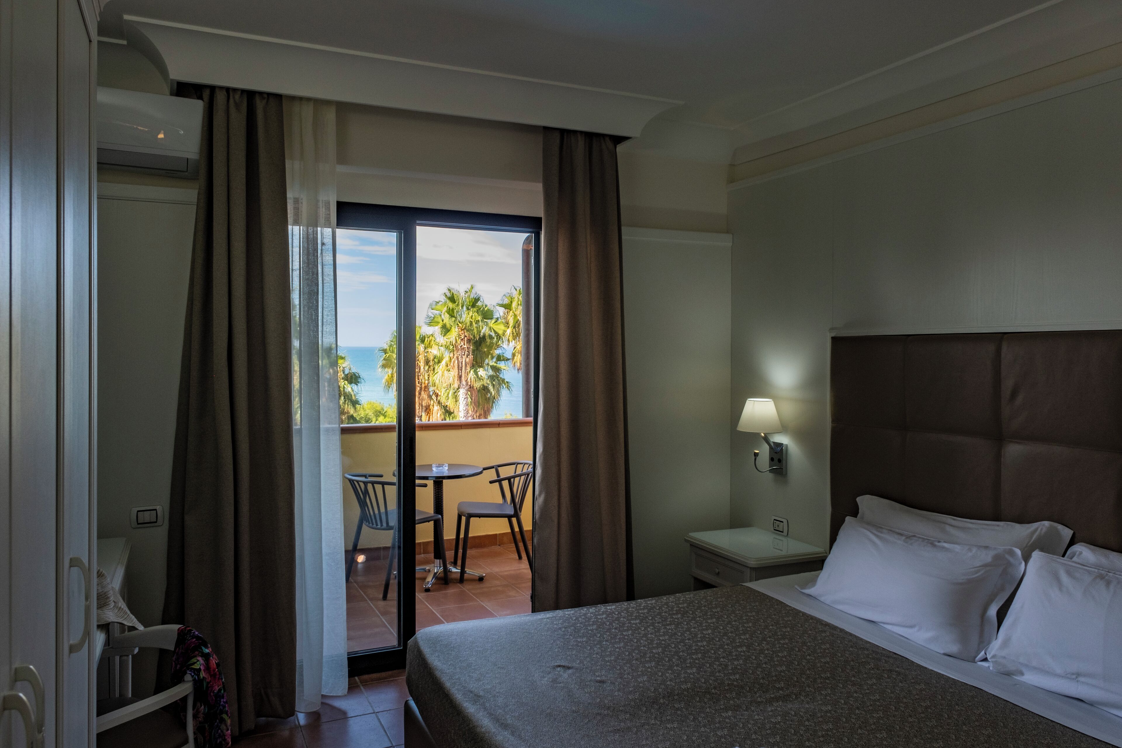 A hotel room with a bed, a nightstand, a lamp, a window with curtains, and a view of palm trees and the ocean.
