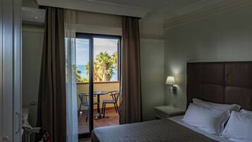 A hotel room with a bed, a nightstand, a lamp, a window with curtains, and a view of palm trees and the ocean.