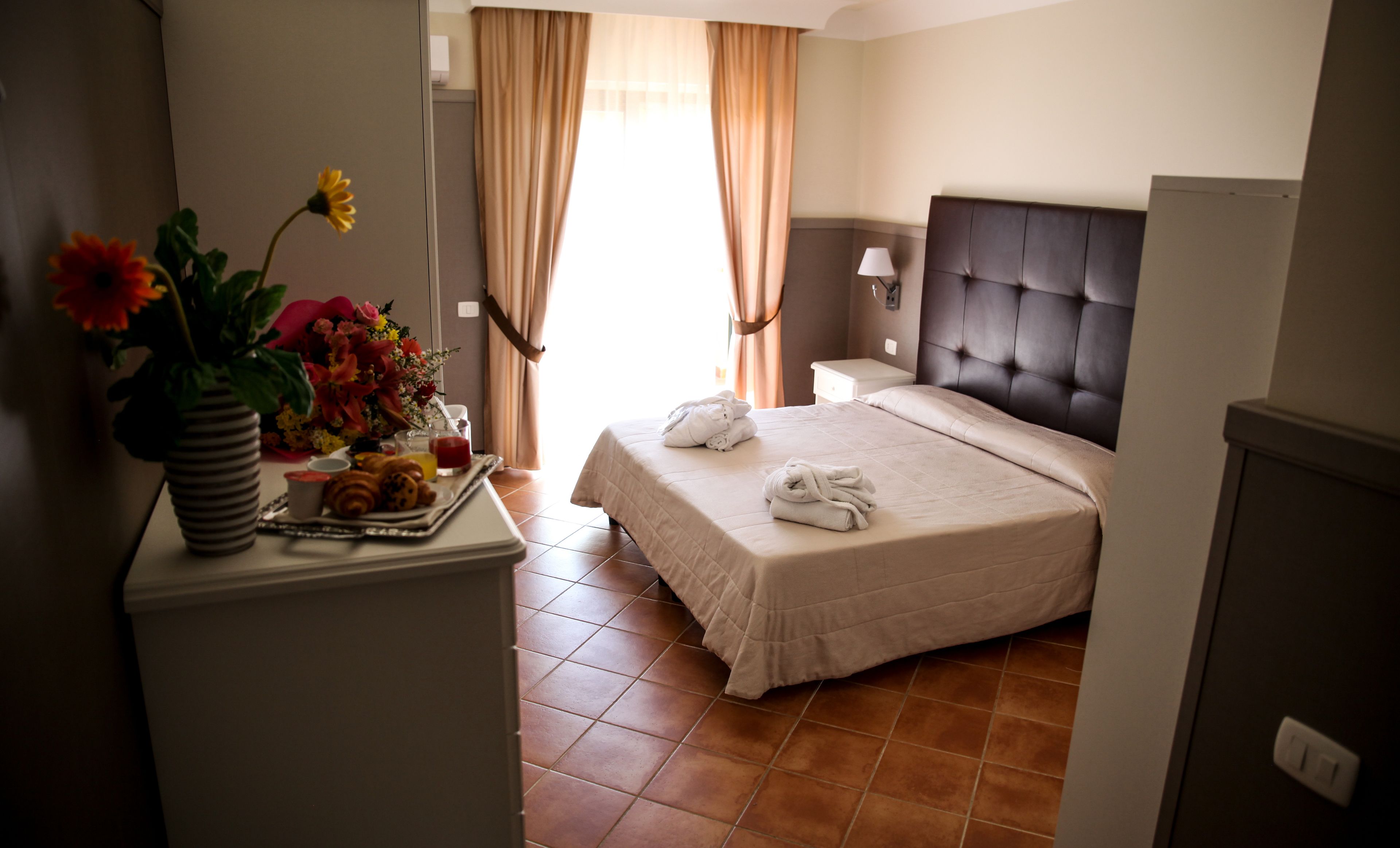 A hotel room with a bed, a bedside table, a lamp, a vase with flowers, and a tray with breakfast items.
