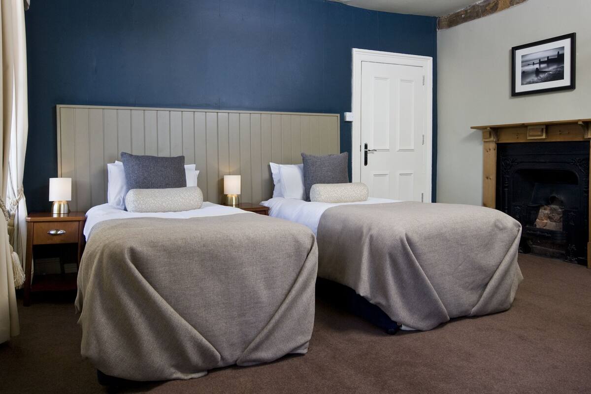A double room with two beds, a nightstand with a lamp, a white door, and a fireplace.