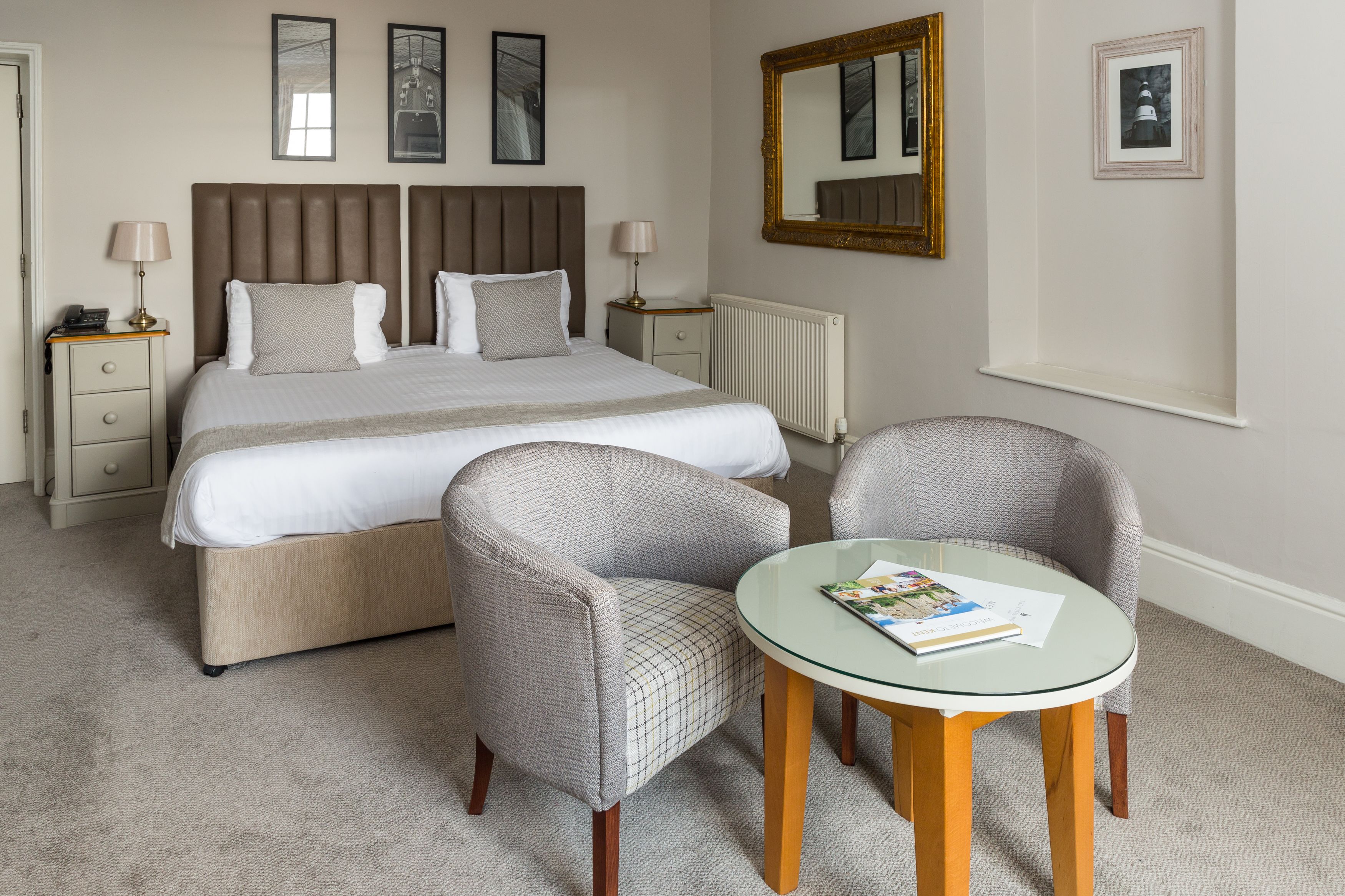 A hotel room with a large bed, two bedside tables, a round glass table, two upholstered chairs, and a large mirror on the wall.