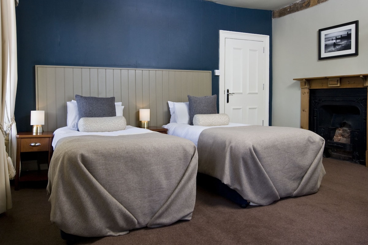 A hotel room with two beds, a nightstand with a lamp, a door, and a fireplace.