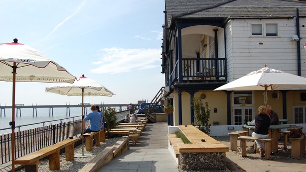 Une terrasse au bord de l’eau, avec des bancs et des tables en bois, des parasols et un bâtiment doté d’un balcon.