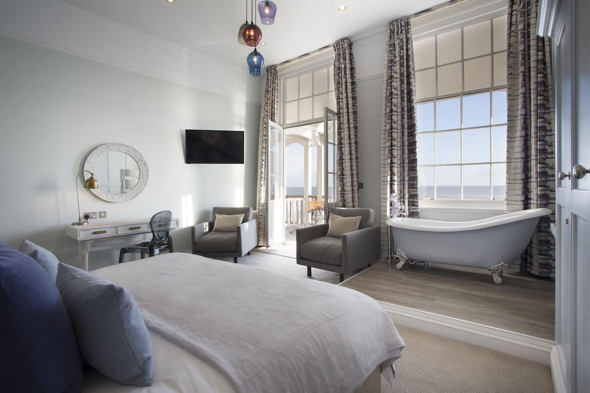 A spacious bedroom with a large bed, a sitting area with chairs, a vanity with a mirror, and a classic clawfoot bathtub.
