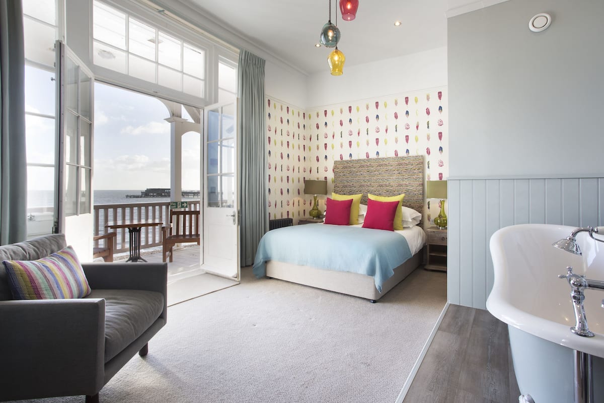 A bedroom with a bed, a sofa, a bathroom with a bathtub, and a view of the sea.