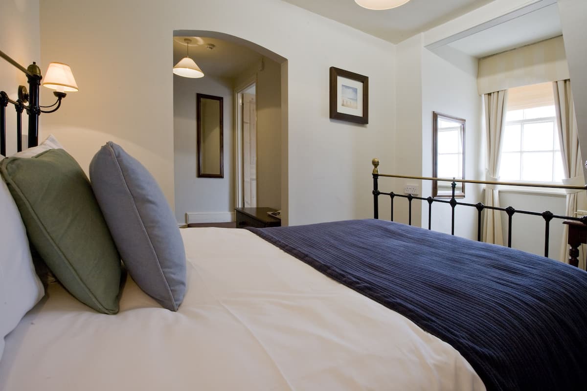 A neatly made bed with a dark blue bedspread and two pillows, a bedside table with a lamp, a framed picture on the wall, and a window with curtains.