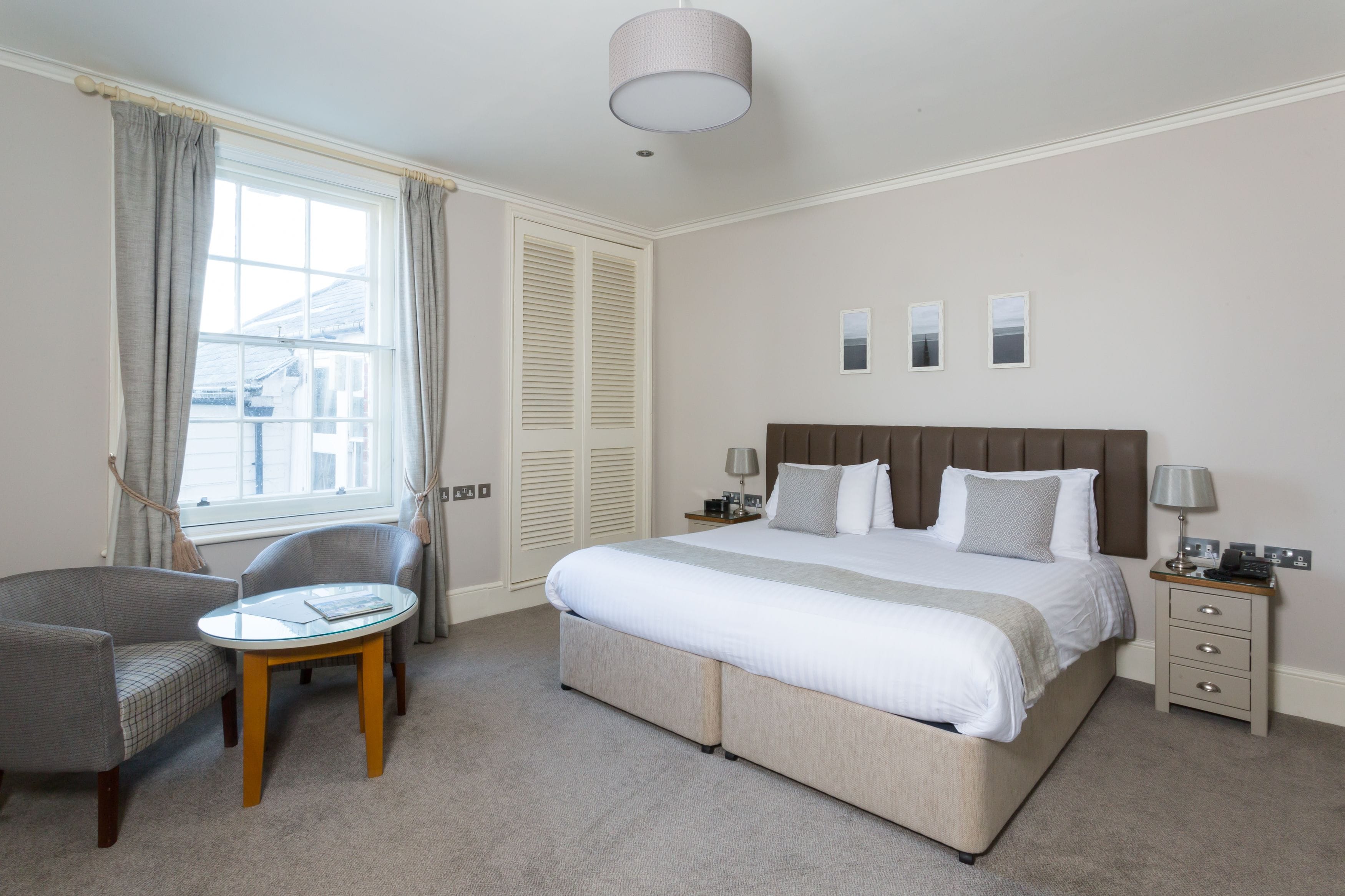 A bedroom with a large bed, a bedside table, a chair, a small round table, and a window with curtains.