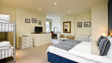 A hotel room with a bunk bed, a desk with a computer, a chair, a bed with a blue accent, and several framed pictures on the walls.
