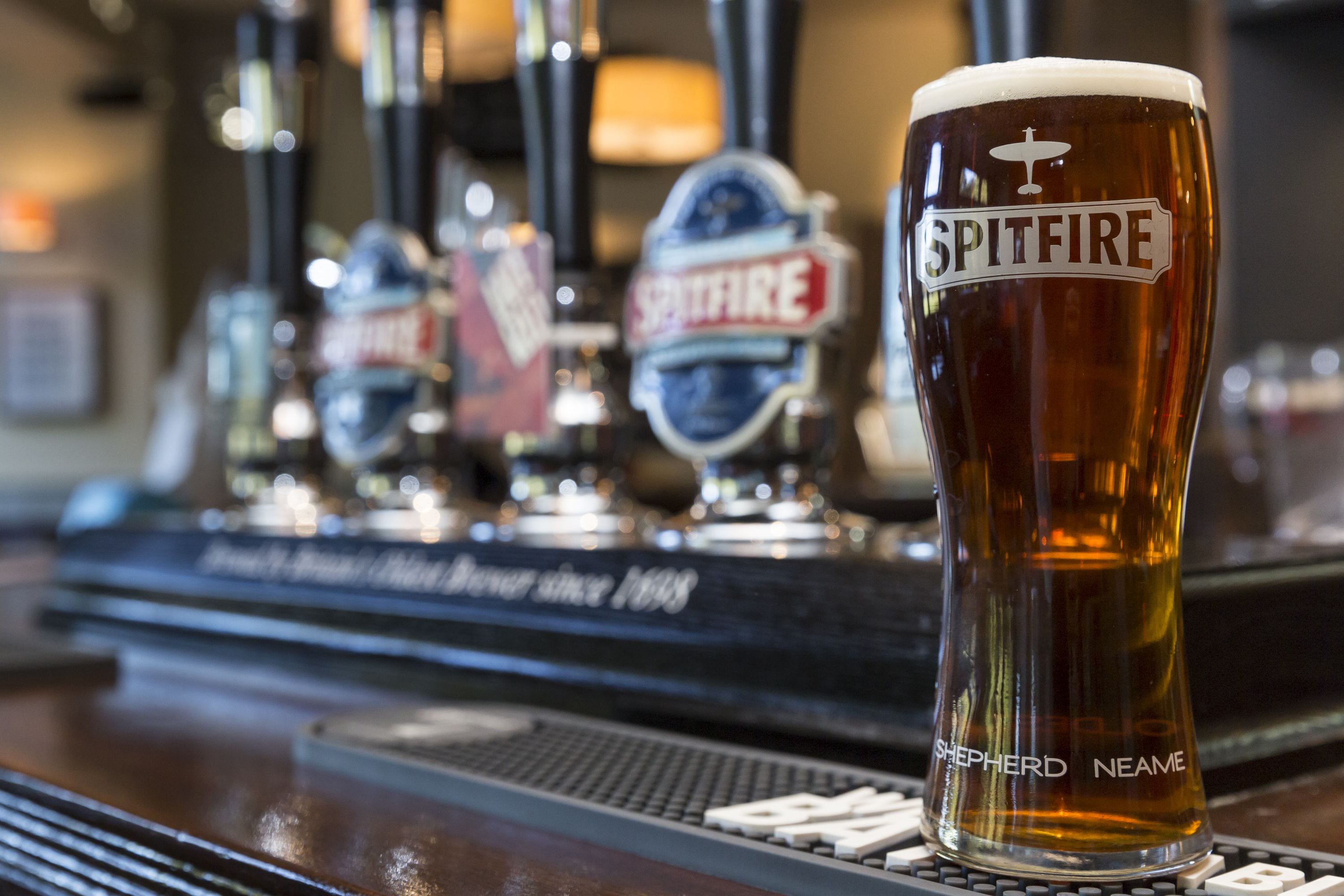 Egy pohár sör, melyen a „SPITFIRE” és a „SHEPHERD NEAME” felirat látható, egy bárpulton, a háttérben pedig sörhordókkal.