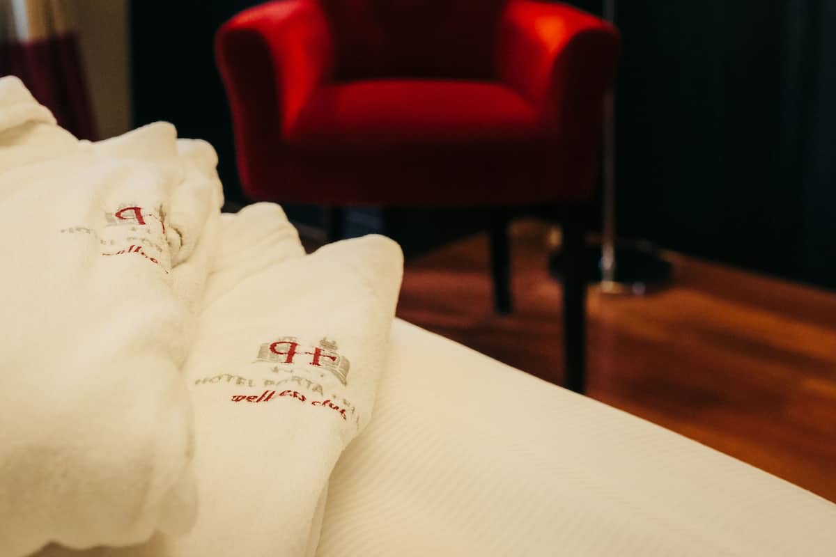 A hotel room with a red chair, a white bed with a blanket, and a floor lamp.