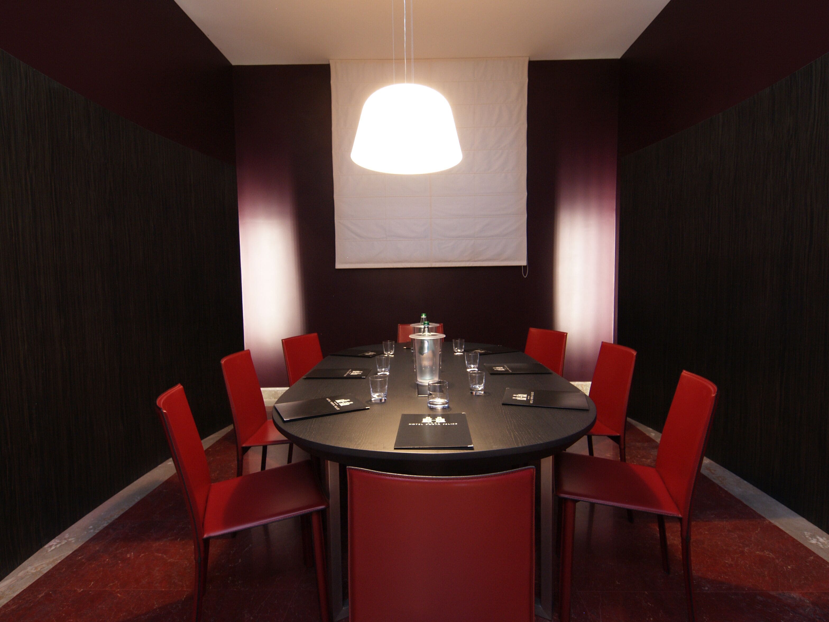 Een vergaderzaal met een ronde tafel, rode stoelen, een projectiescherm en een hanglamp.