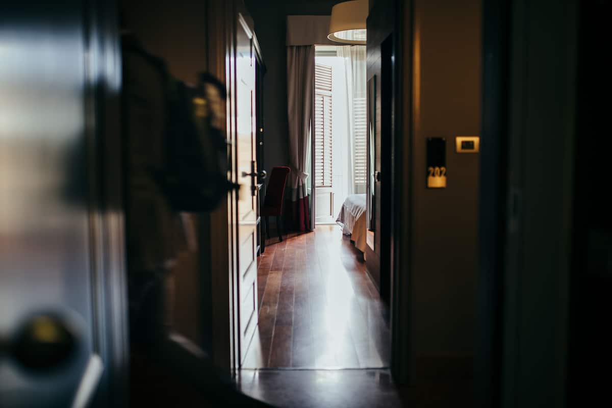 A hotel hallway with wooden flooring, a bed with a white bedspread, and a door with a number plate.