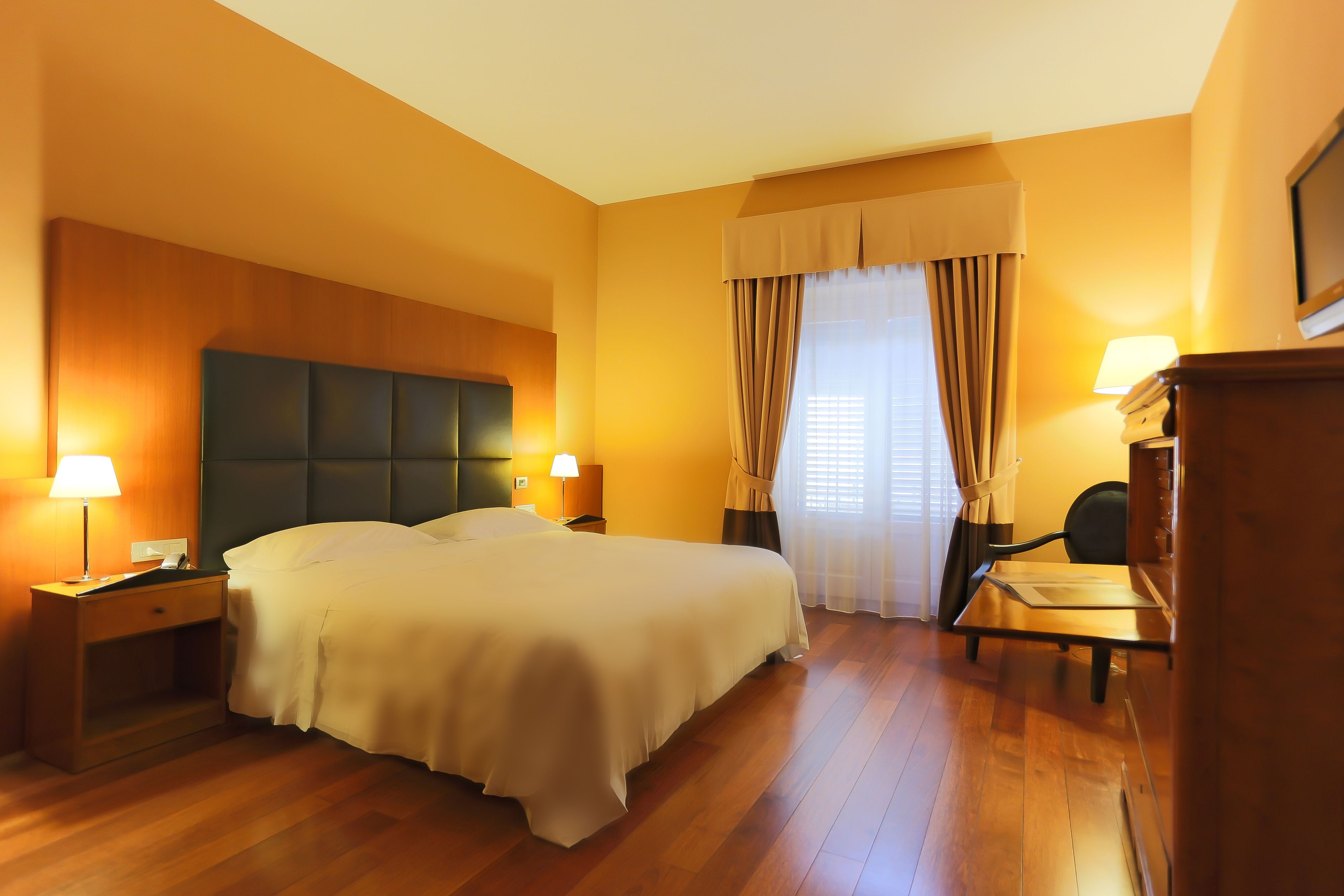 A hotel room with a large bed, two bedside tables with lamps, a wooden headboard, a desk with a chair, and a window with curtains.