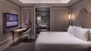 Quarto de hotel com uma cama grande, uma escrivaninha com cadeira, televisão, banheiro com espelho e pia, e uma TV na parede.