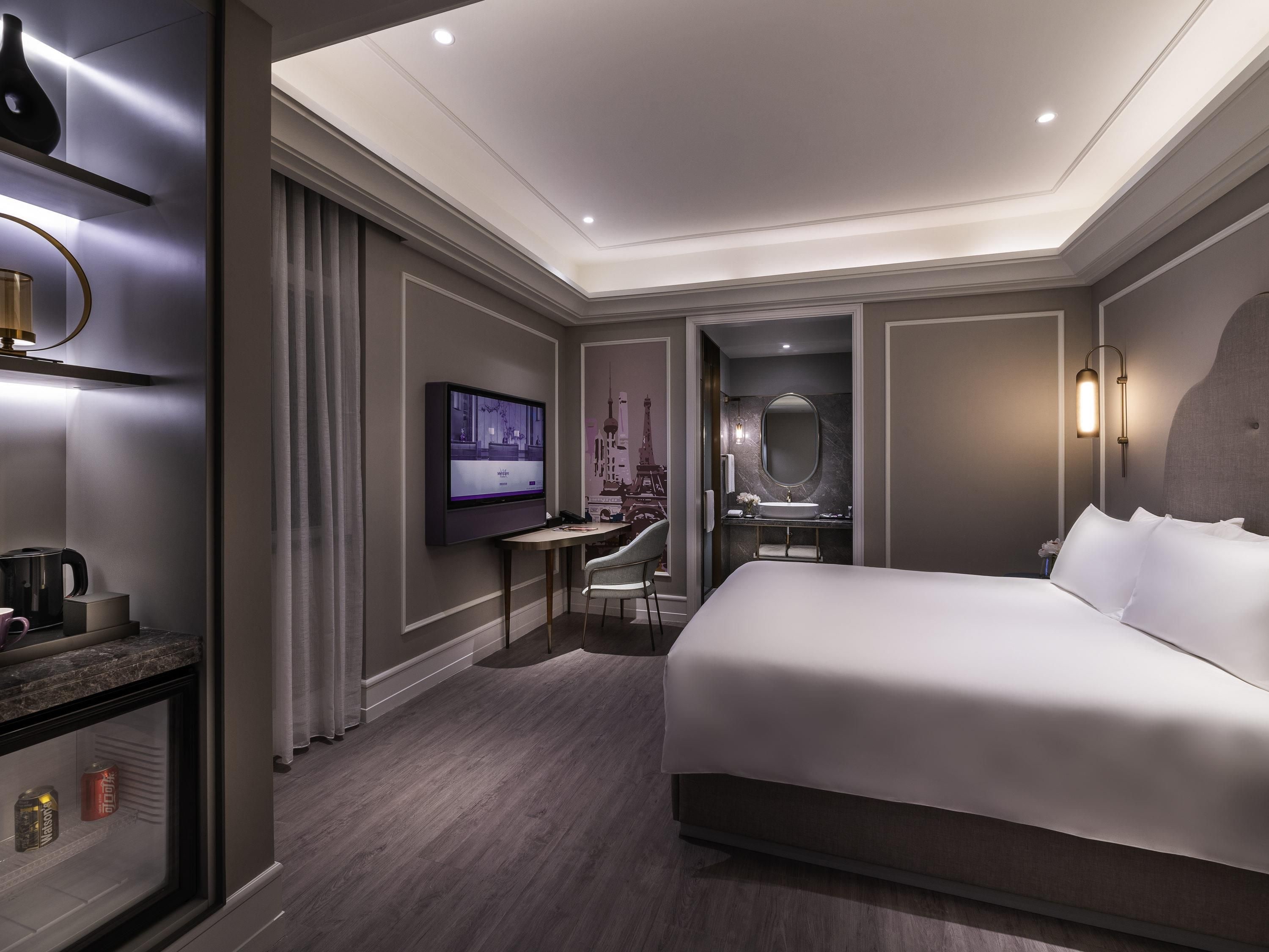A modern hotel room with a large bed, a television, a desk with a chair, a bathroom with a mirror, and a decorative Eiffel Tower on the wall.