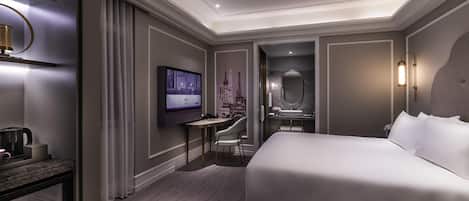 A modern hotel room with a large bed, a television, a desk with a chair, a bathroom with a mirror, and a decorative Eiffel Tower on the wall.