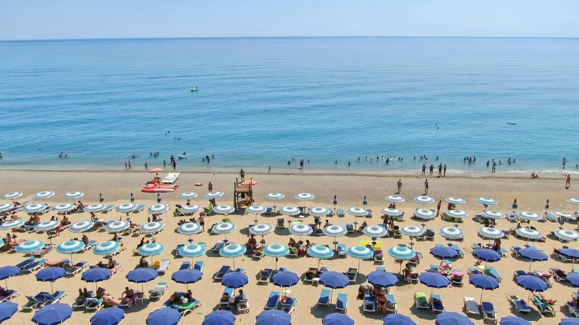 Une plage avec de nombreux parasols et des chaises longues, une tour de surveillance pour les sauveteurs, et l’océan en arrière-plan.