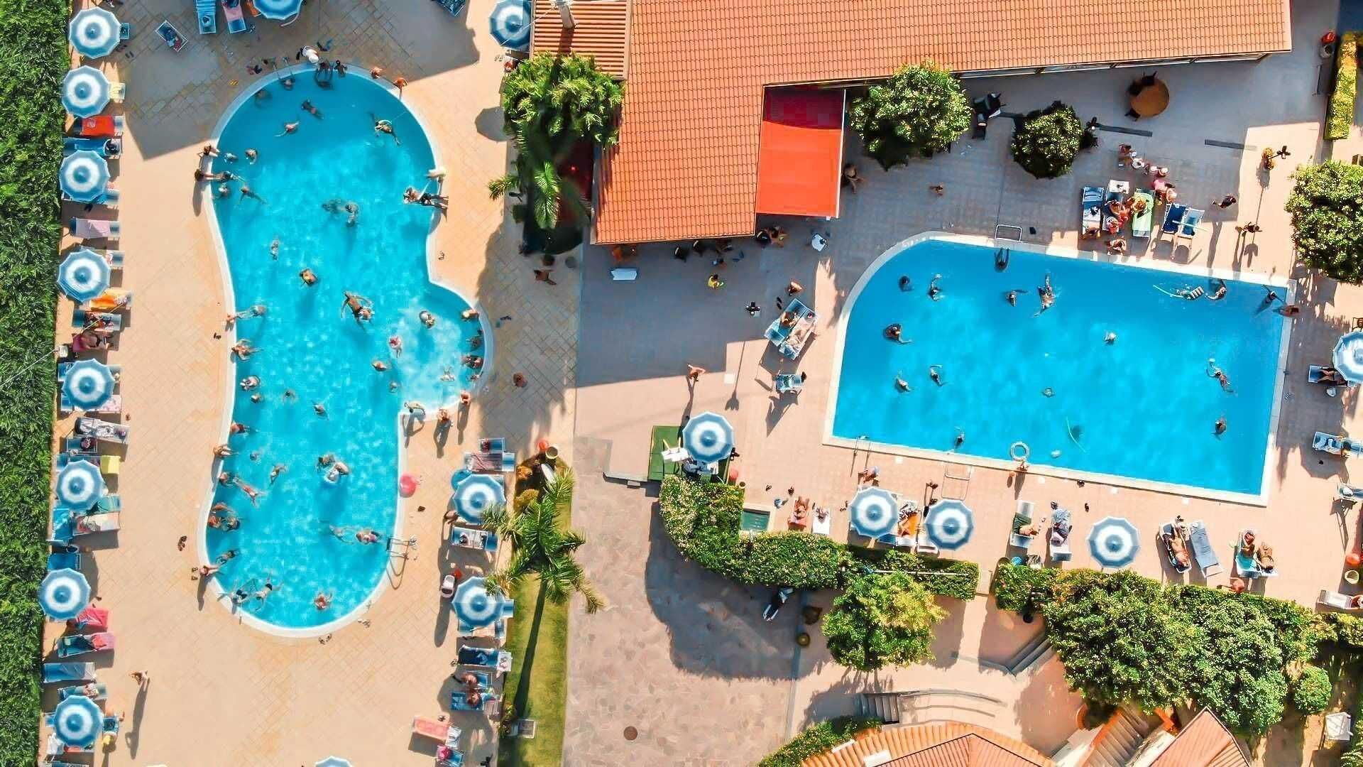 Vista aérea de un resort con dos piscinas grandes rodeadas de tumbonas y sombrillas, un área cubierta con techo rojo y un pequeño edificio.