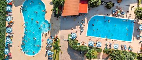 Vista aérea de un resort con dos piscinas grandes rodeadas de tumbonas y sombrillas, un área cubierta con techo rojo y un pequeño edificio.