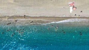 Vue aérienne d’une plage aux eaux turquoise et limpides, avec une tour de surveillance et des personnes qui se baignent et font de la planche à voile.