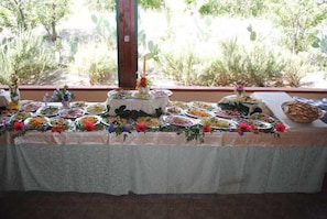 Una mesa dispuesta con diversos platos y un pastel, decorada con flores, en una habitación con vistas a cactus y árboles.