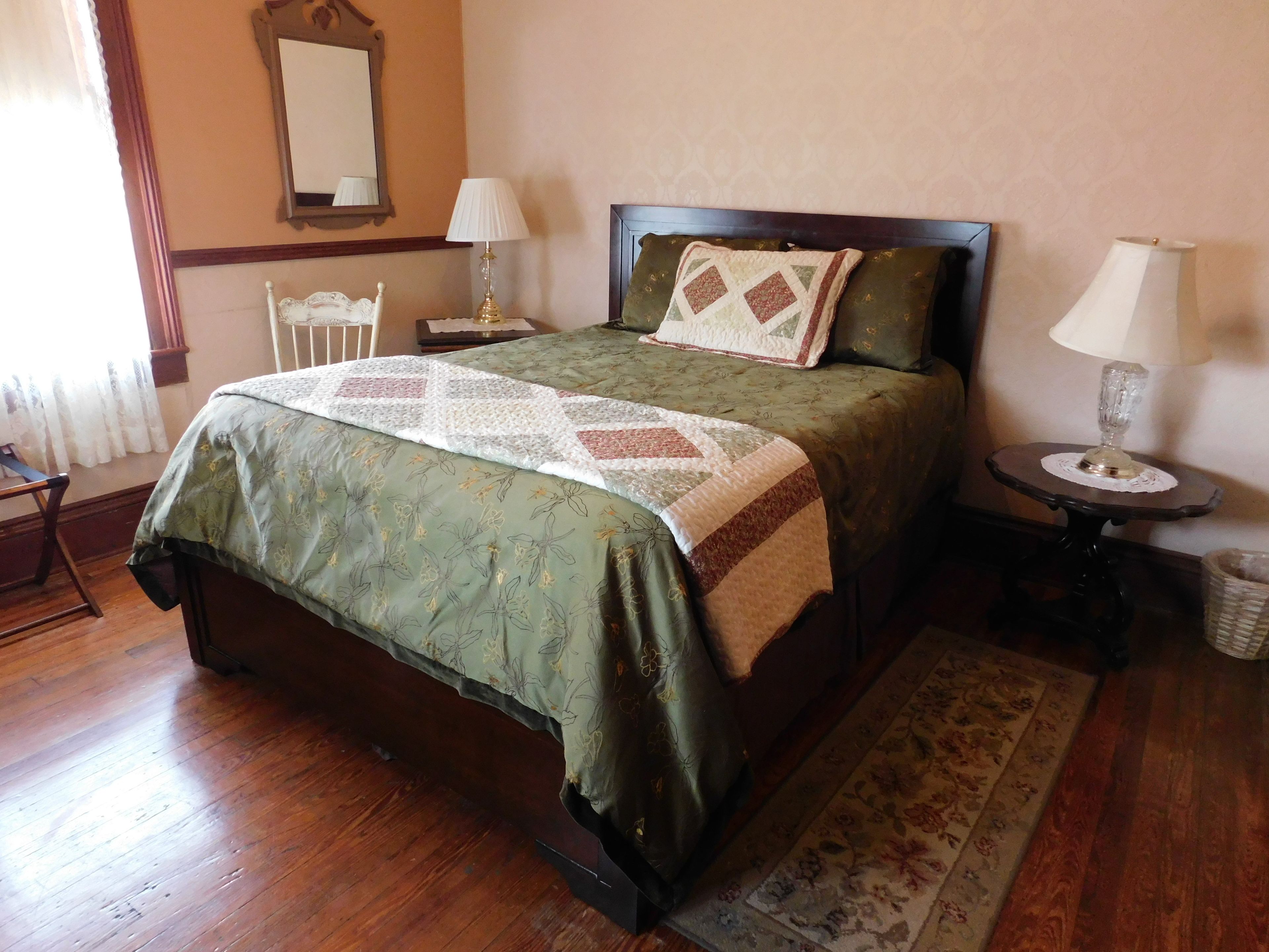 Una cama bien tendida con cubrecama verde y colcha, una mesita de noche con lámpara, una silla y una mesita redonda pequeña con lámpara.