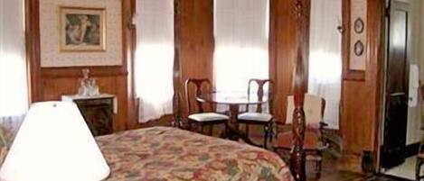 Una habitación con una cama, una mesa y dos sillas. Hay una lámpara sobre la cama, un cuadro en la pared y una puerta al fondo.