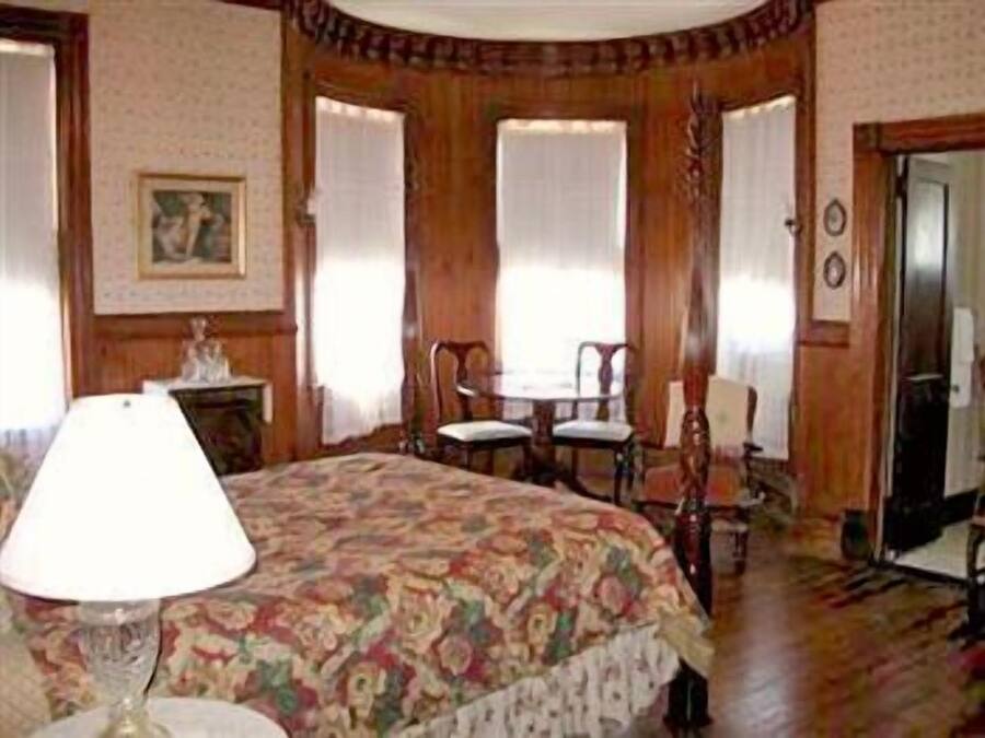 Une pièce comprenant un lit, une table et deux chaises. On y trouve une lampe sur le lit, un tableau au mur et une porte au fond.