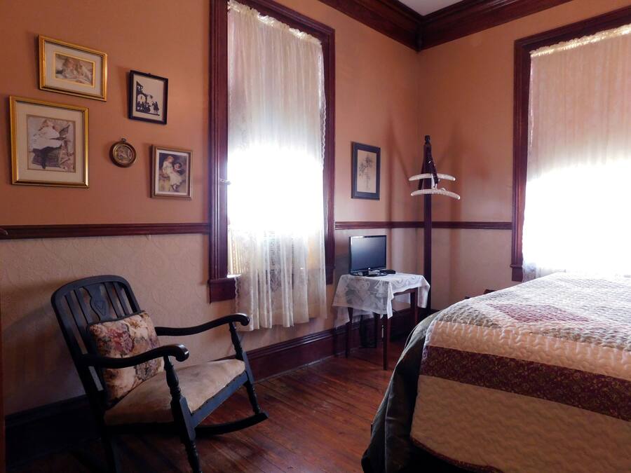 Quarto com piso de madeira, uma cadeira de balanço, uma cama com colcha, uma escrivaninha com computador e quadros nas paredes.