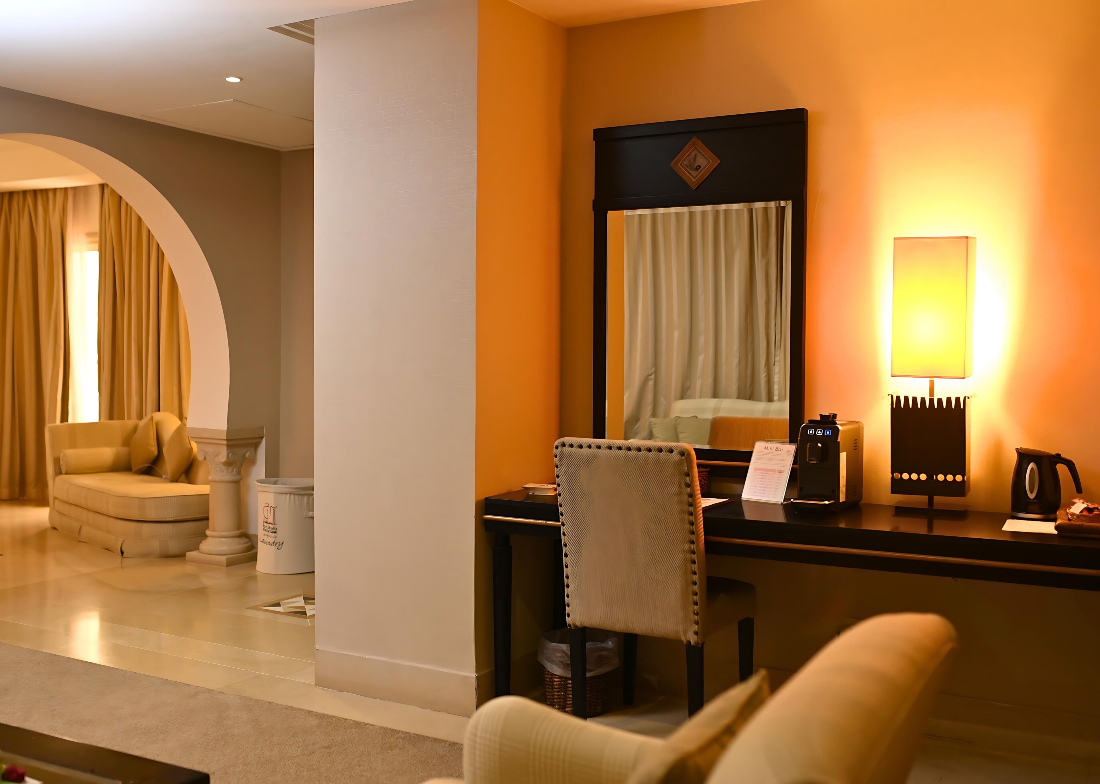 Habitación de hotel con sofá, una silla, escritorio con lámpara, cafetera y minibar.