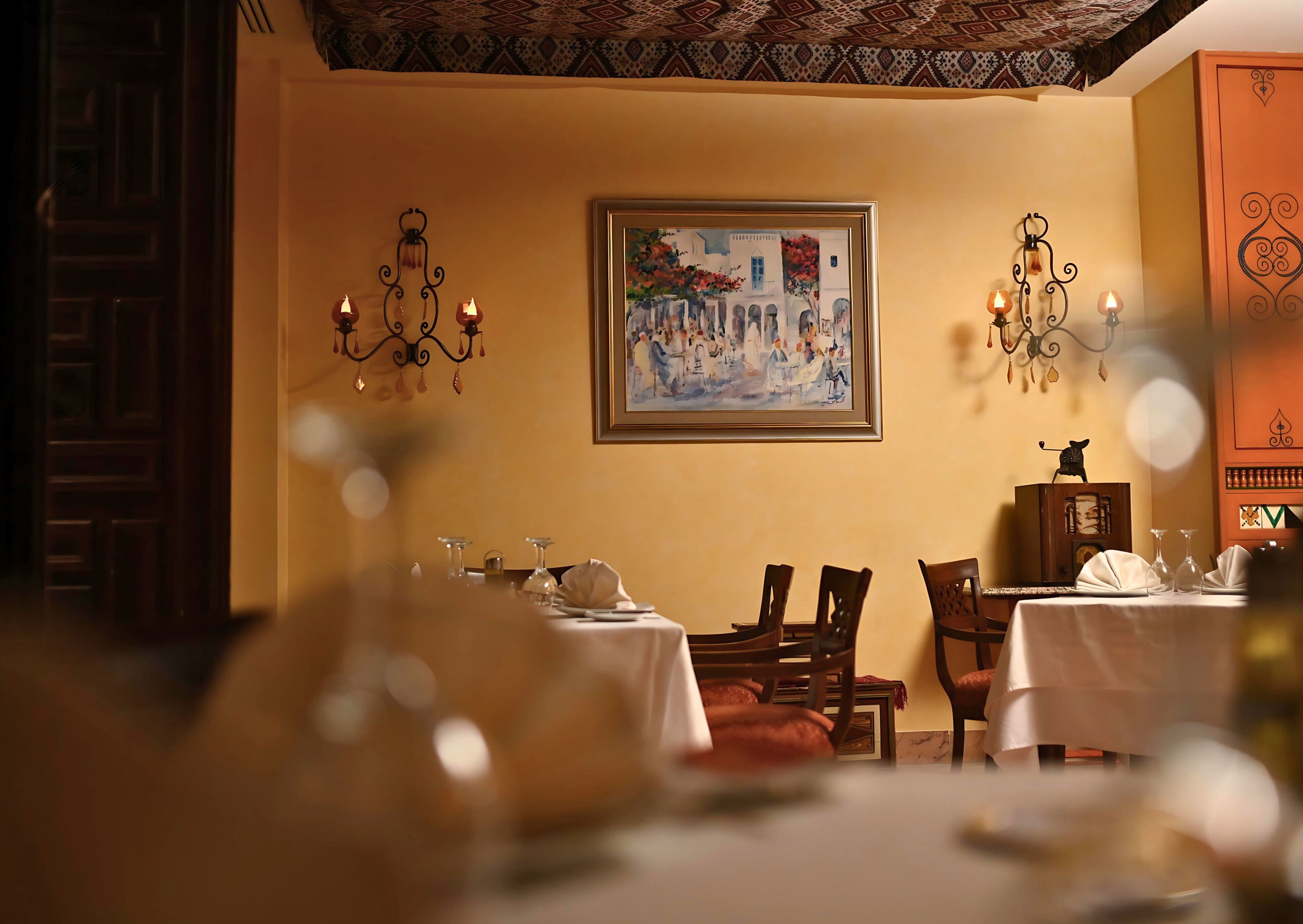 A dining area with a table set for two, a painting on the wall, and a decorative cabinet.