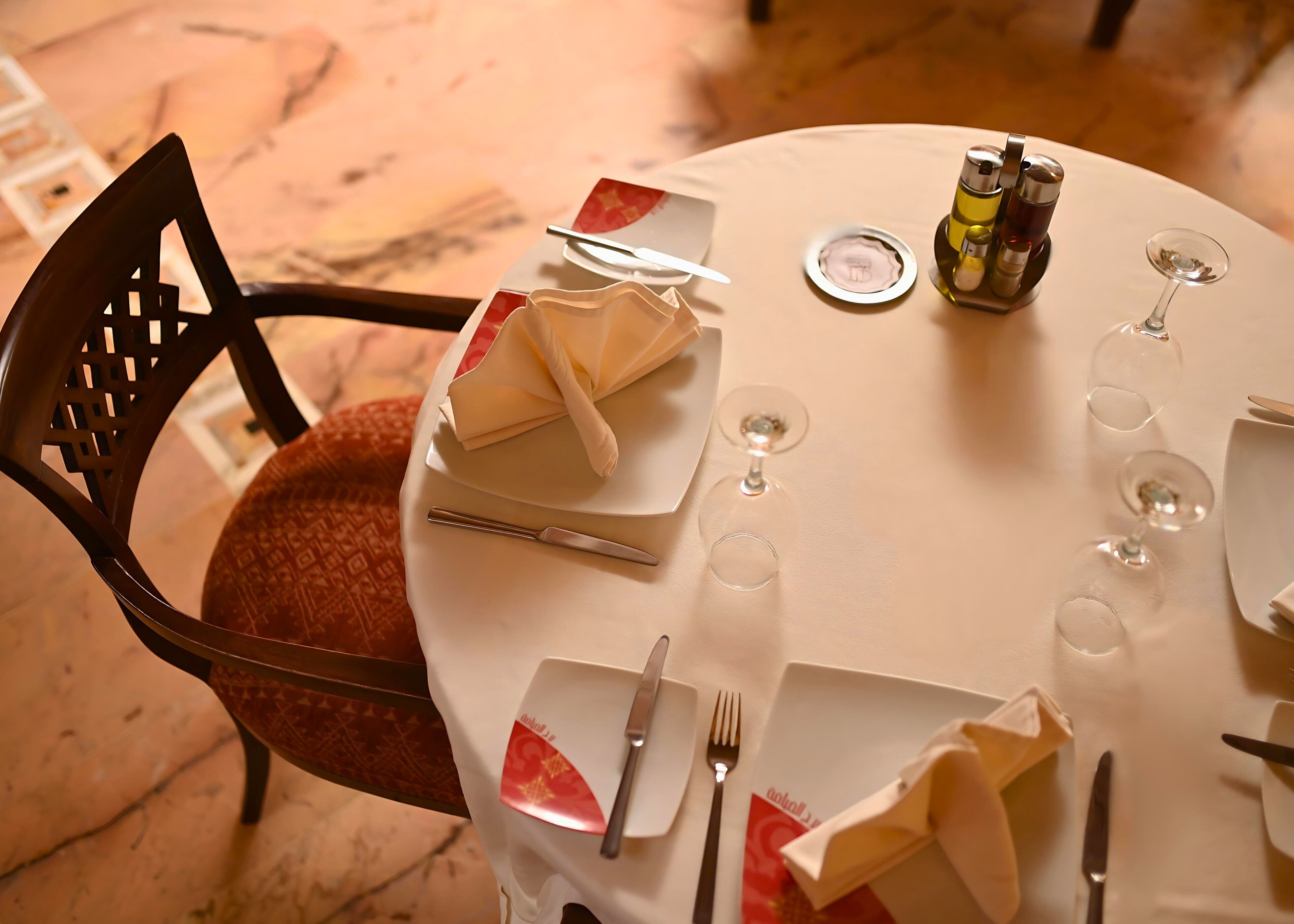 A round dining table with a white tablecloth, neatly set with plates, cutlery, and wine glasses.