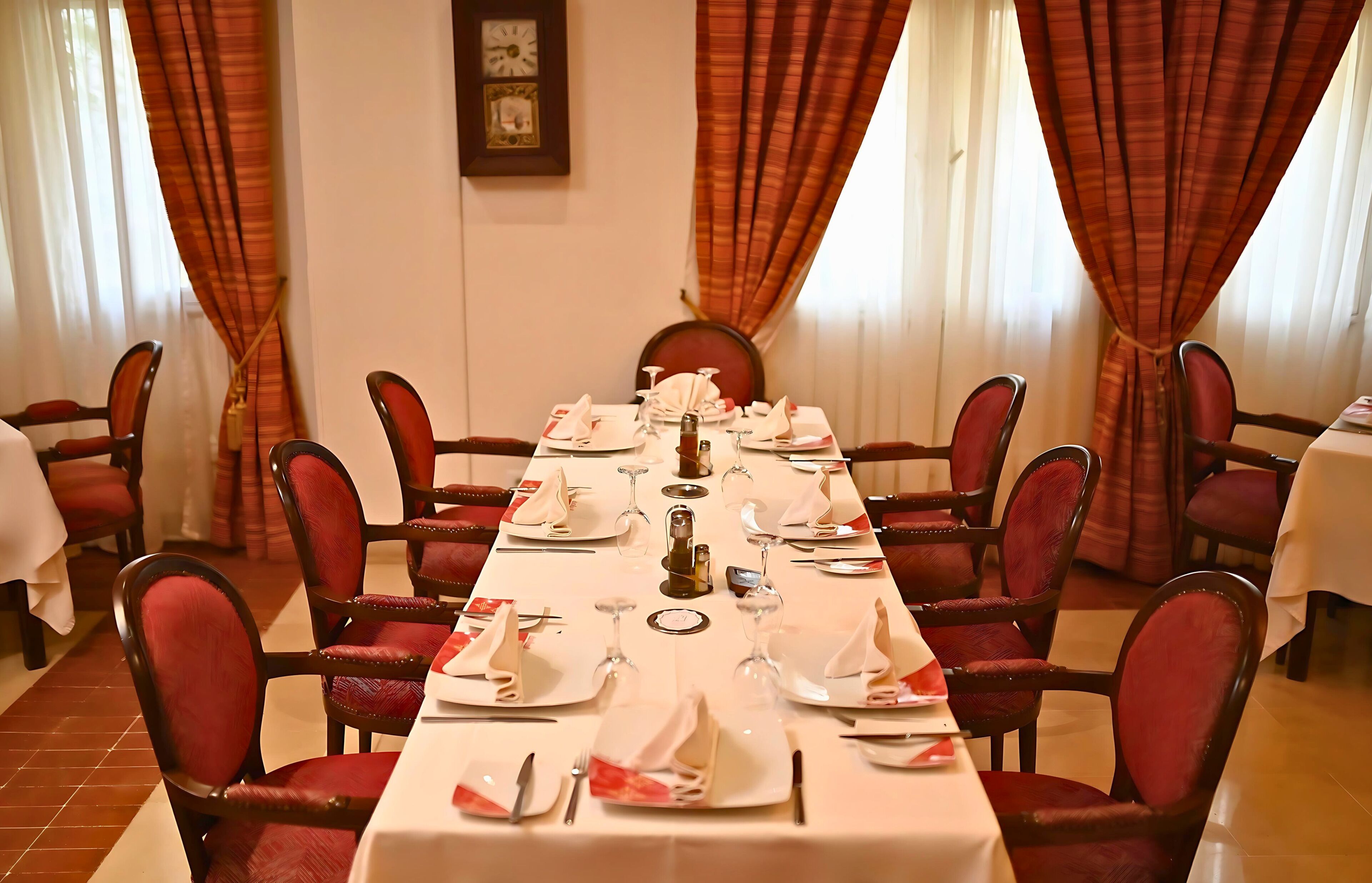 Zona de dinar amb una taula llarga preparada per a dinar, envoltada de cadires tapissades de vermell.