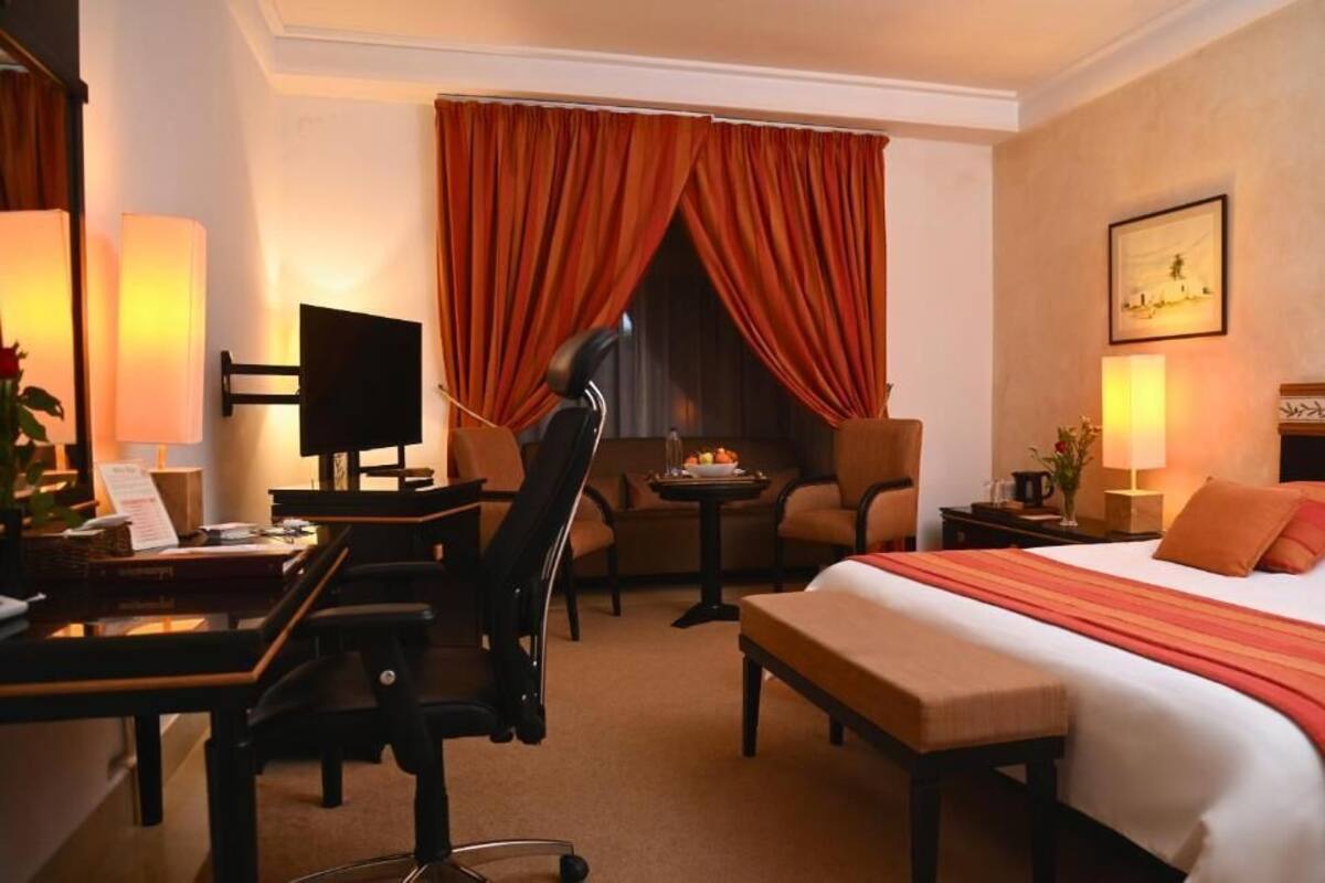 A hotel room with a bed, desk, chair, TV, and a small table with a lamp.