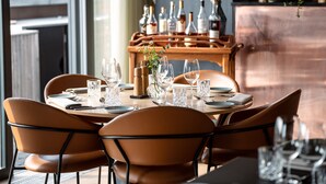 A dining area with a round table set for four, surrounded by leather chairs. A wooden sideboard with bottles and glasses is visible in the background.