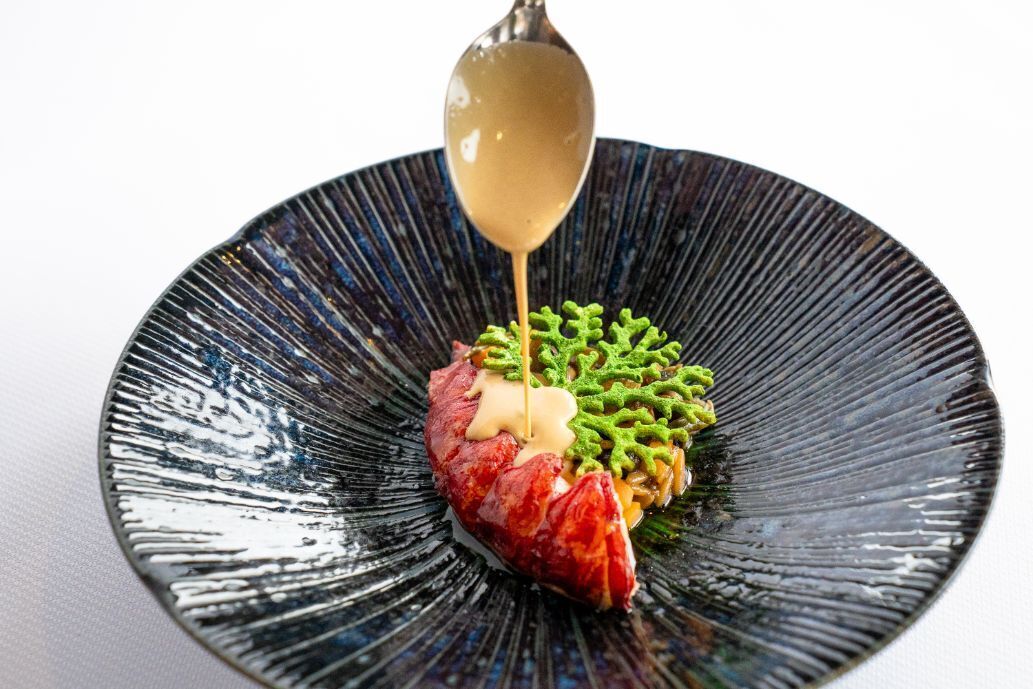 A black plate with a spoonful of sauce being poured onto a piece of meat and green garnish.