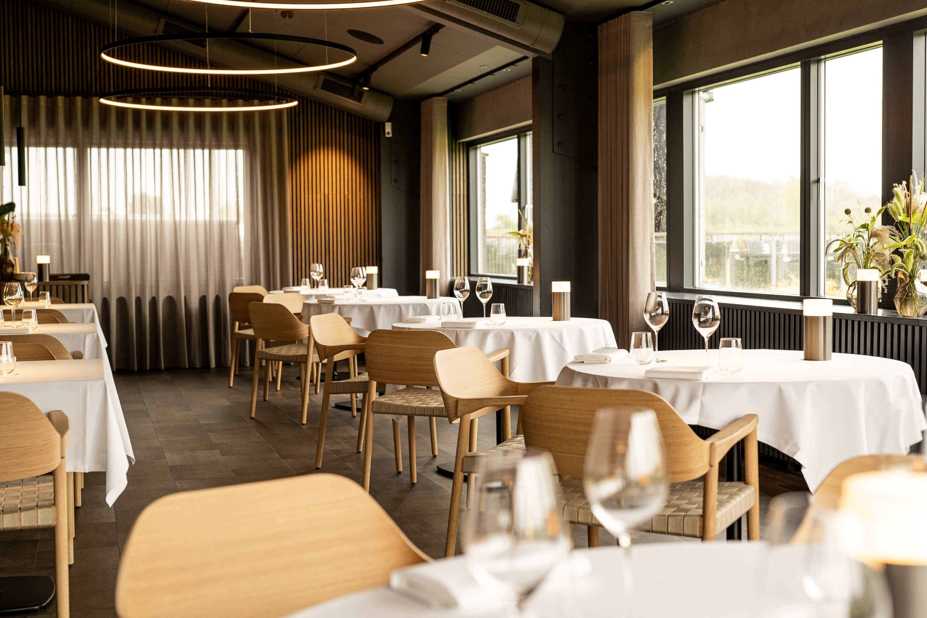 En restaurant med flere borde og stole, store vinduer og et moderne interiør.