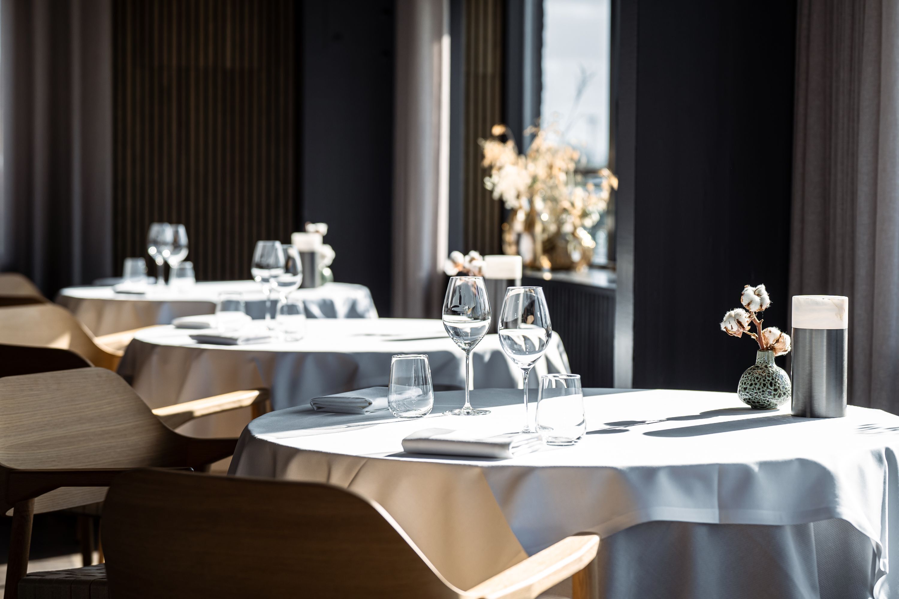 L’intérieur d’un restaurant présente des tables rondes dressées pour le service, avec un design minimaliste aux couleurs neutres et baigné de lumière naturelle.