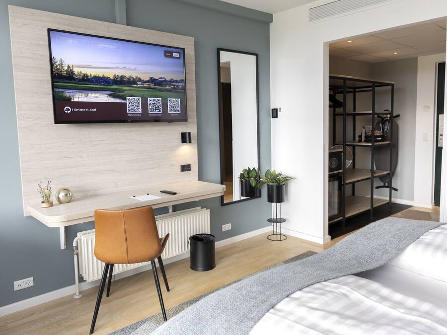 Una habitación de hotel moderna con un televisor grande montado en la pared, un escritorio con una silla, una cama con un tapete gris y una estantería con plantas.