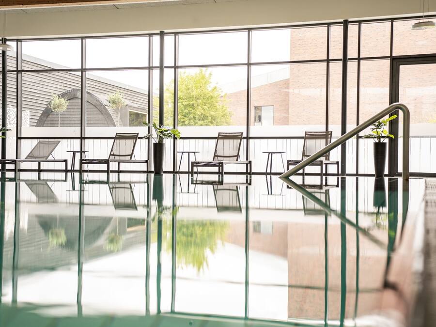 Una piscina interior con paredes de vidrio transparente, sillas de descanso y plantas en macetas.