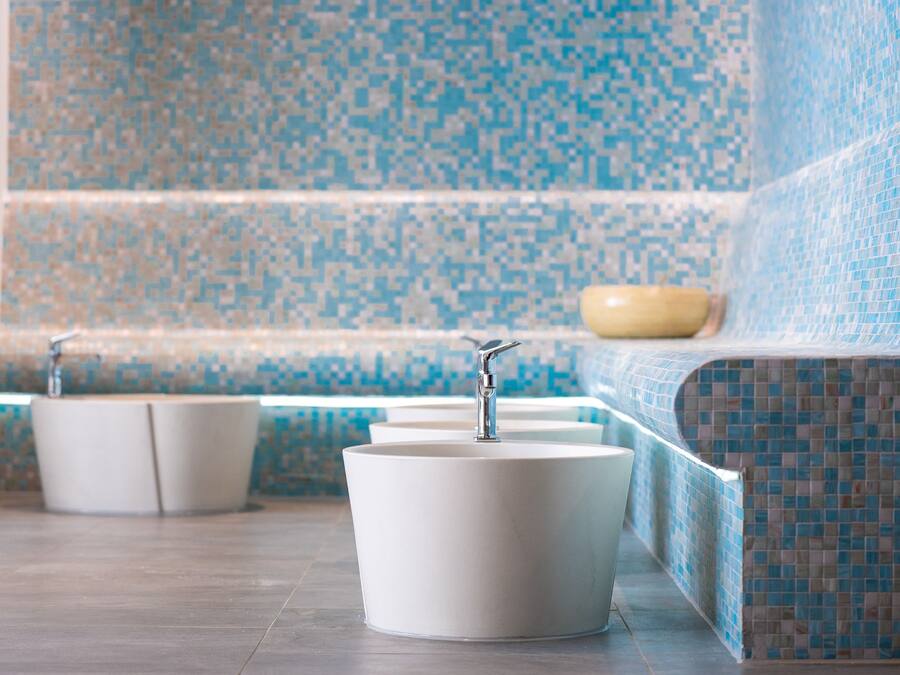 Un baño con tres lavamanos blancos, un cuenco color beige y una pared con mosaico azul.