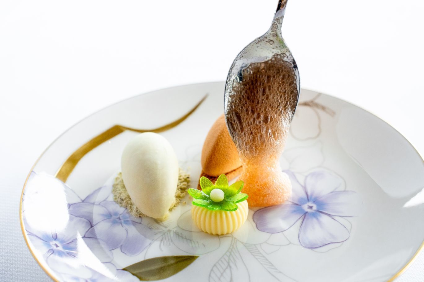 A dessert plate with a white floral pattern, featuring a scoop of ice cream, a piece of cake, and a decorative item with a green garnish.