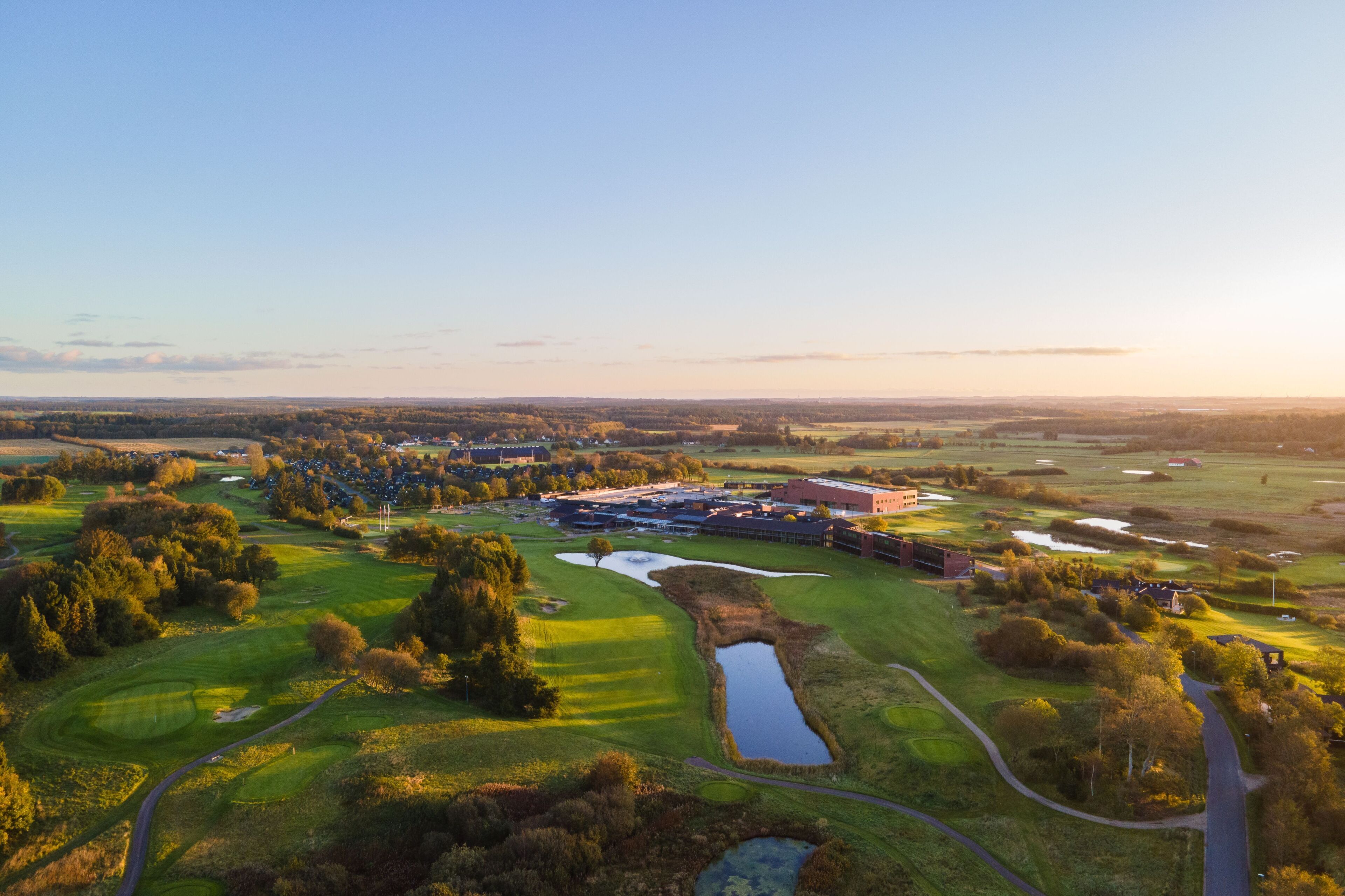 An aerial view of a golf course with a clubhouse, ponds, and surrounding greenery.